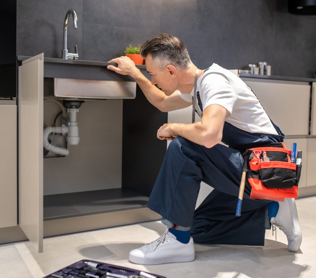 Man kneeling down touching sink inspecting pipes Household repair. Middle-aged man in blue overalls knelt down with hand touching sink carefully examining pipes in kitchen at home
